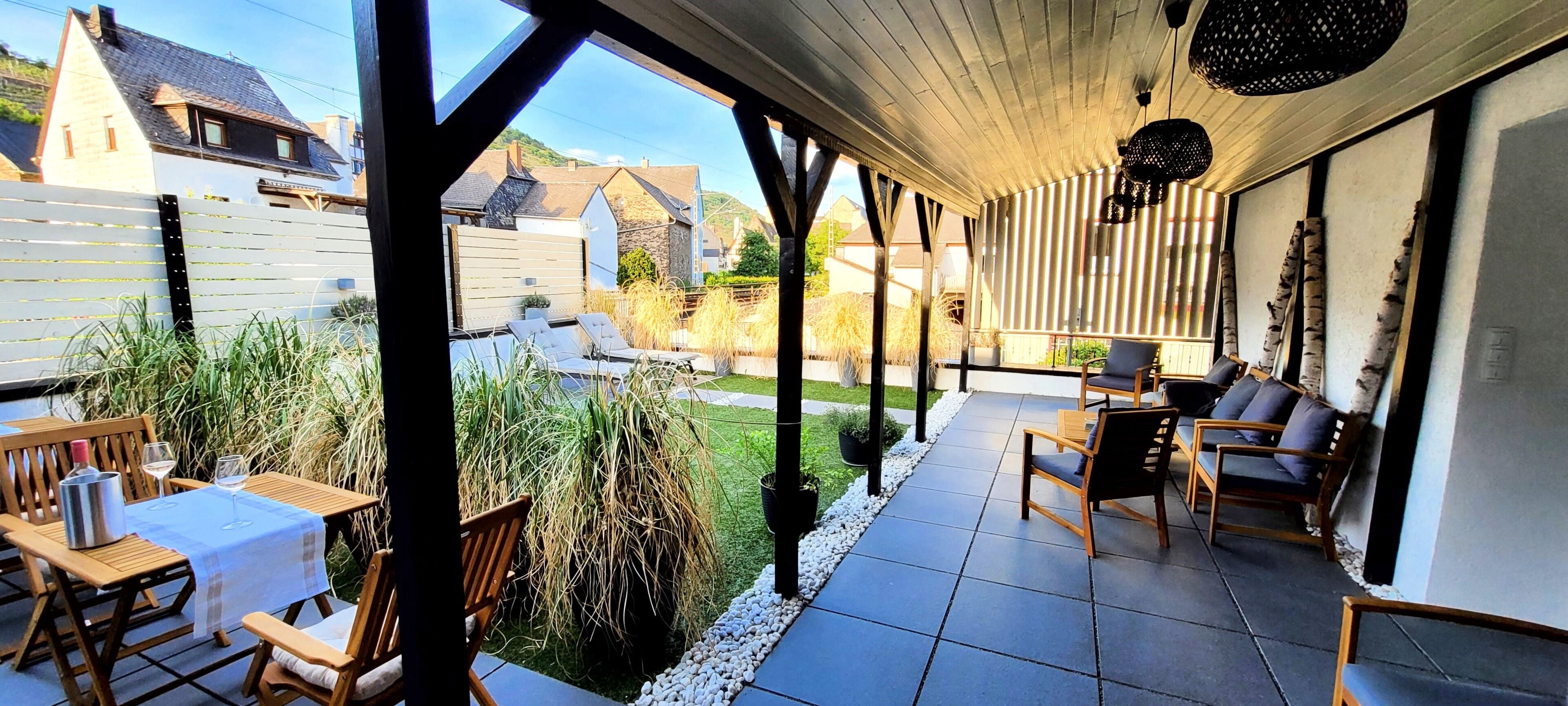 A covered patio with wooden furniture, a table with a white tablecloth, and a view of a garden and neighboring houses.