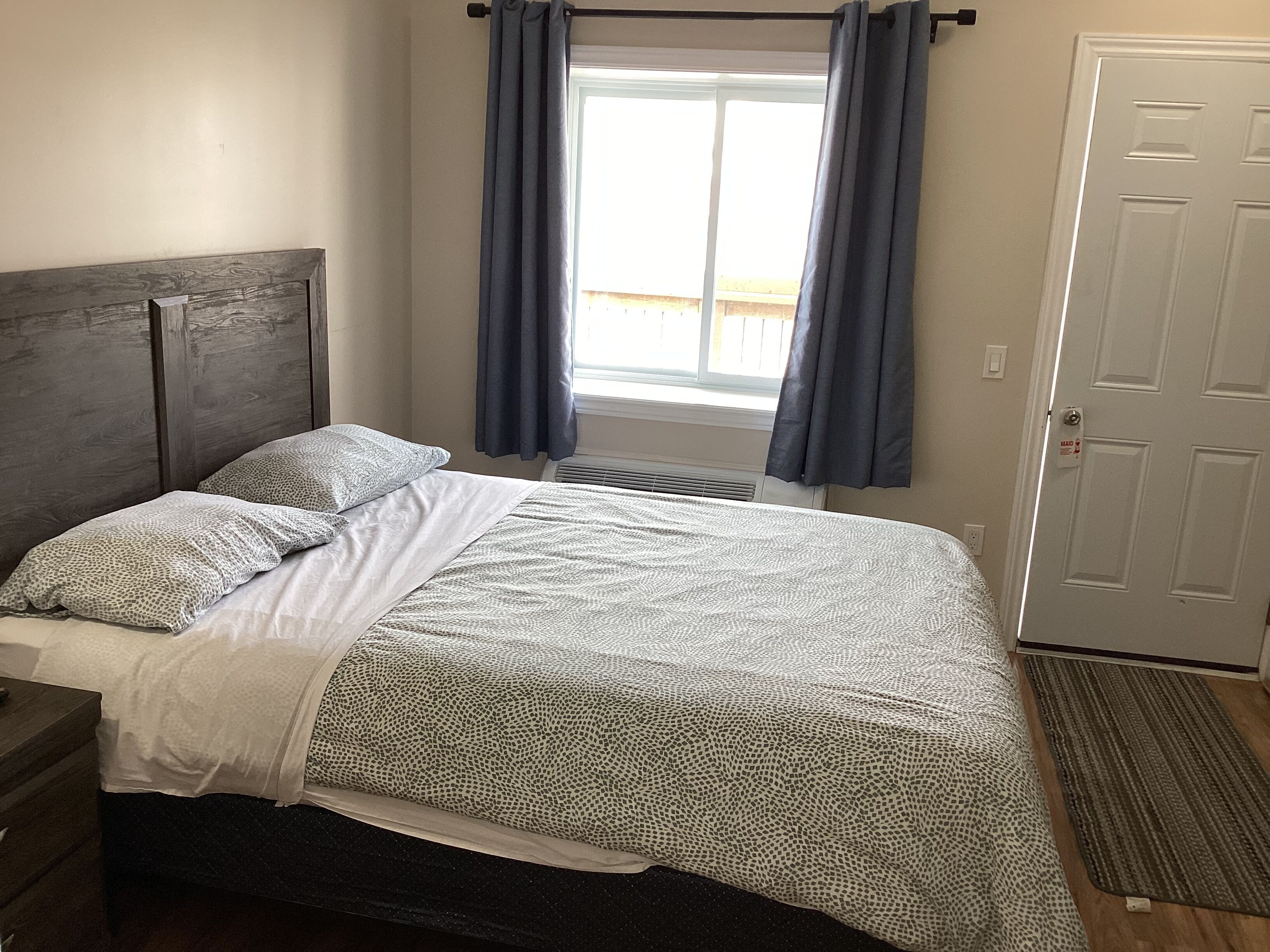 Um quarto com uma cama, cabeceira, janela com cortinas, porta e radiador.