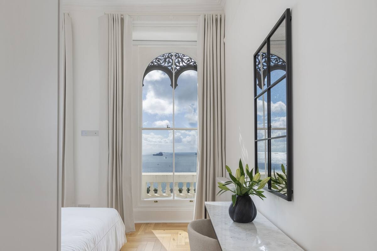 Quarto de hotel estreito com uma cama, uma mesa, uma cadeira e vista para o mar pela janela.