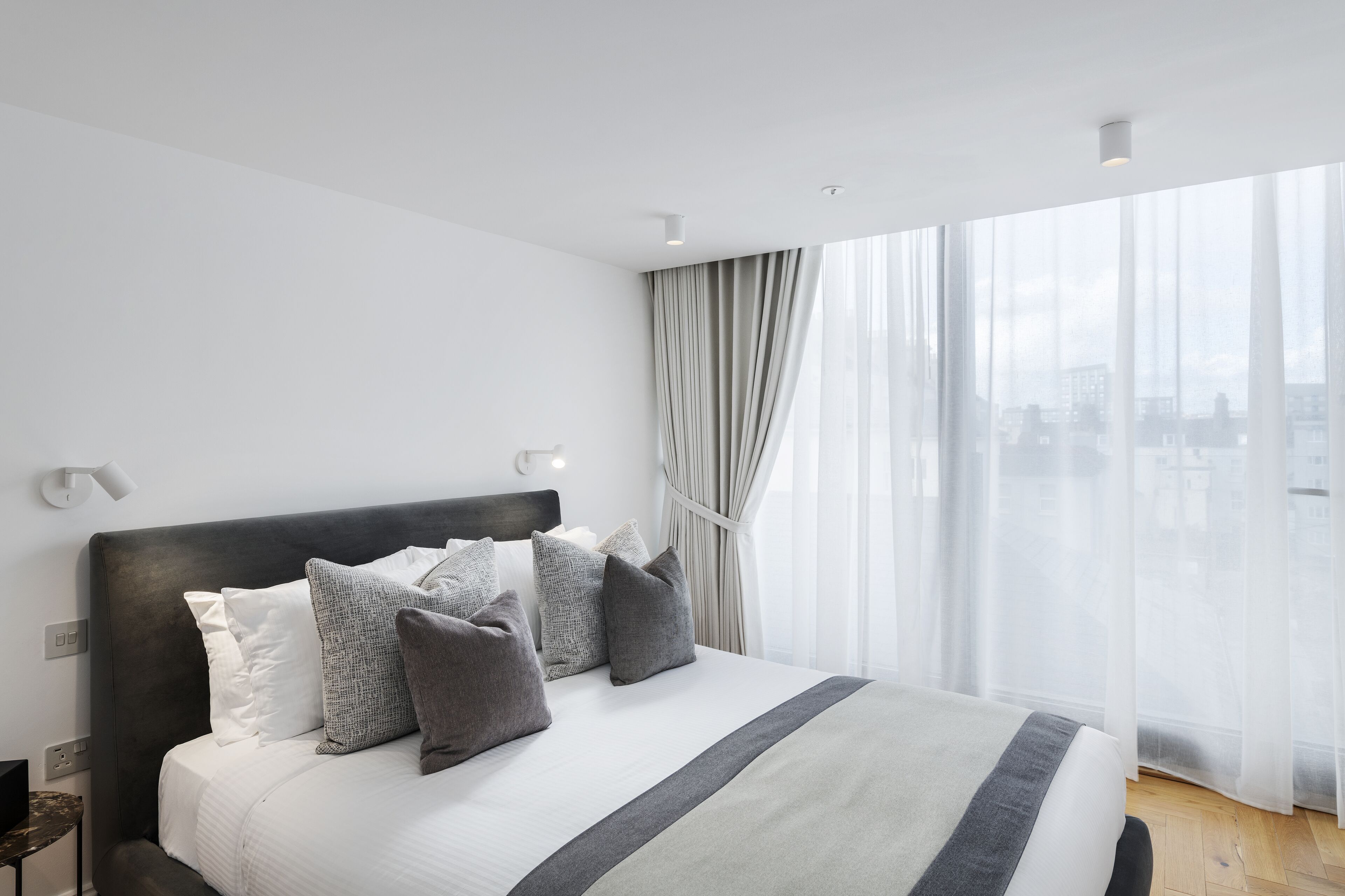 A modern hotel room with a large bed, a city view through sheer curtains, and a minimalist design.