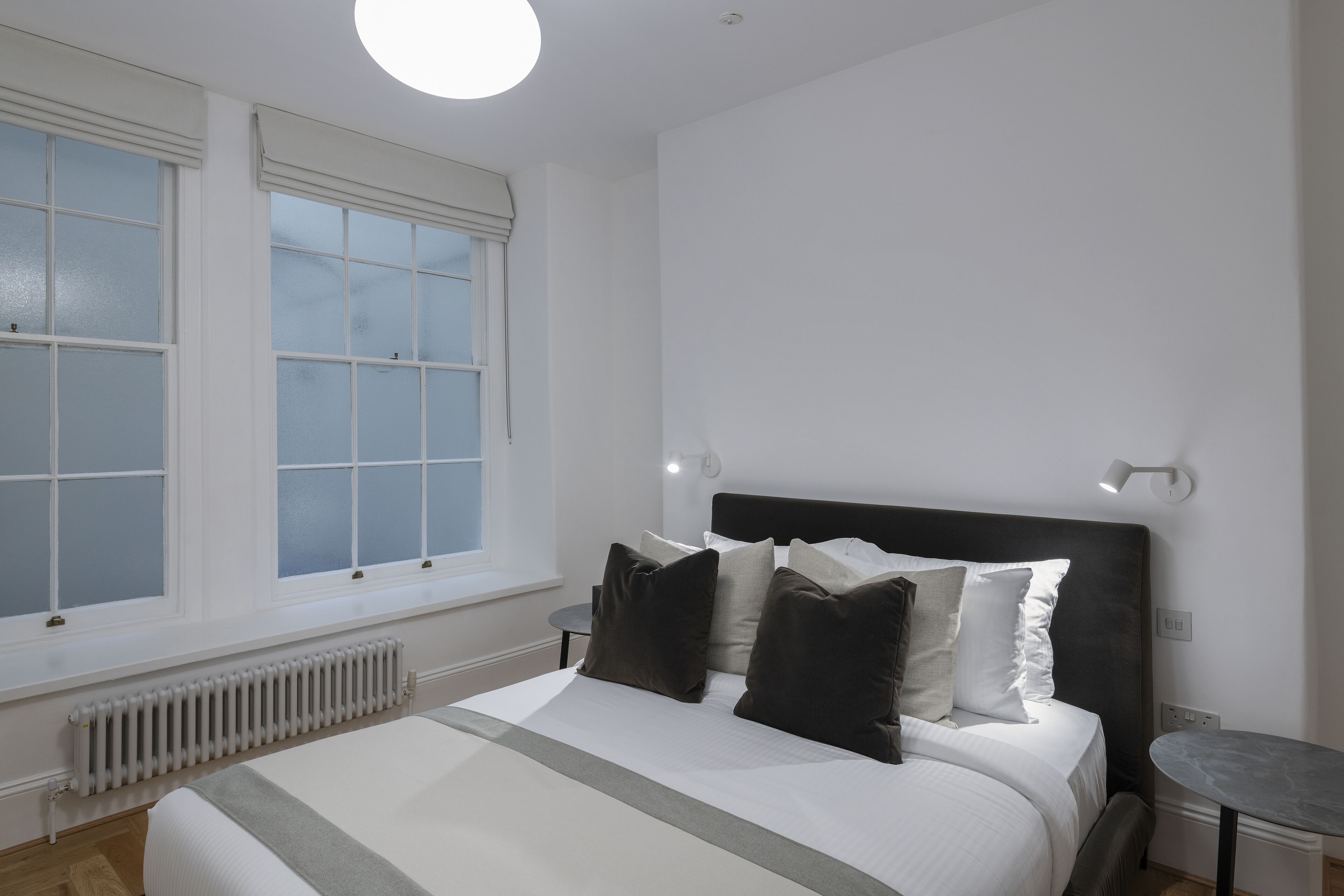 A bedroom with a large bed, white walls, a window, a radiator, and a small table.