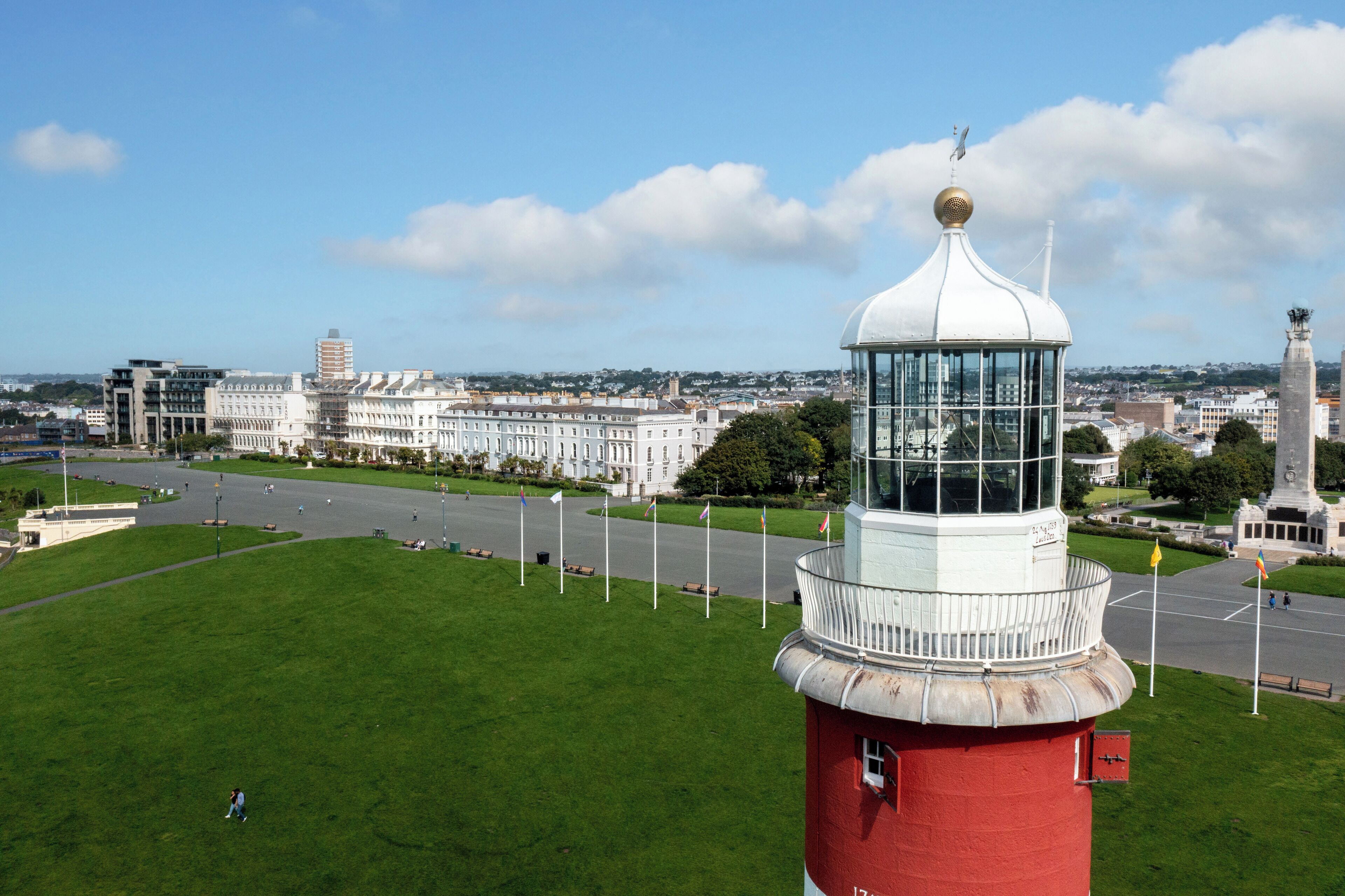 A lighthouse with a red and white structure, a grassy area in the foreground, and a cityscape with buildings in the background.