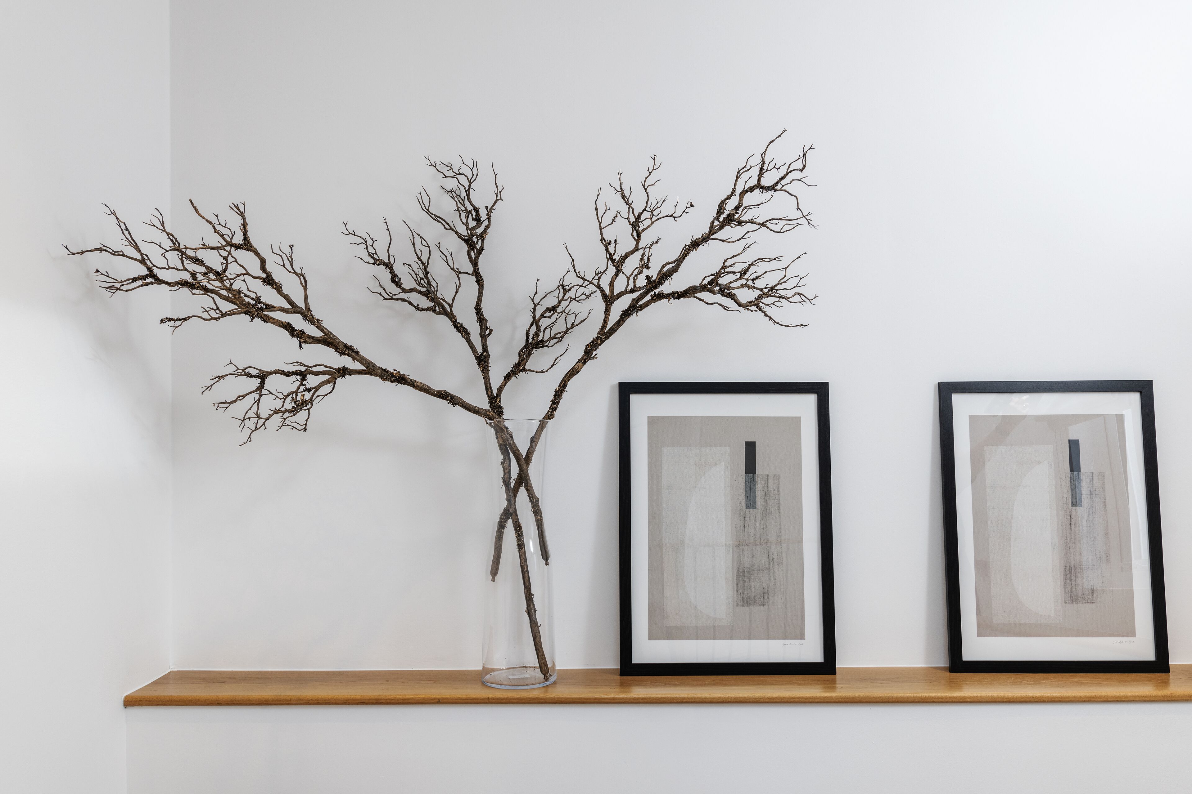 A branch in a clear vase on a shelf with two framed abstract paintings.