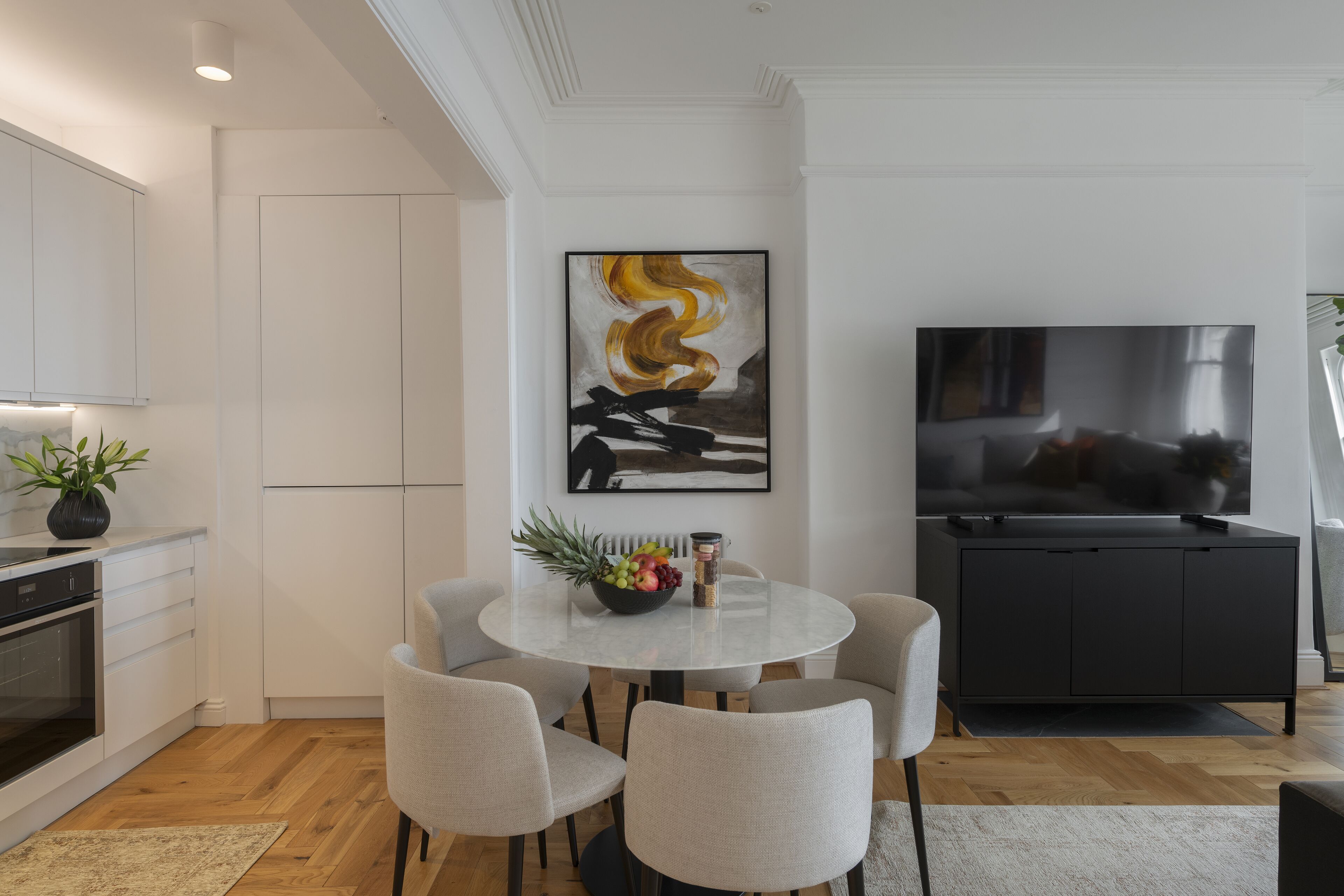 A modern kitchen with white cabinets, a dining area with a round table and chairs, a black cabinet, and a flat-screen TV.