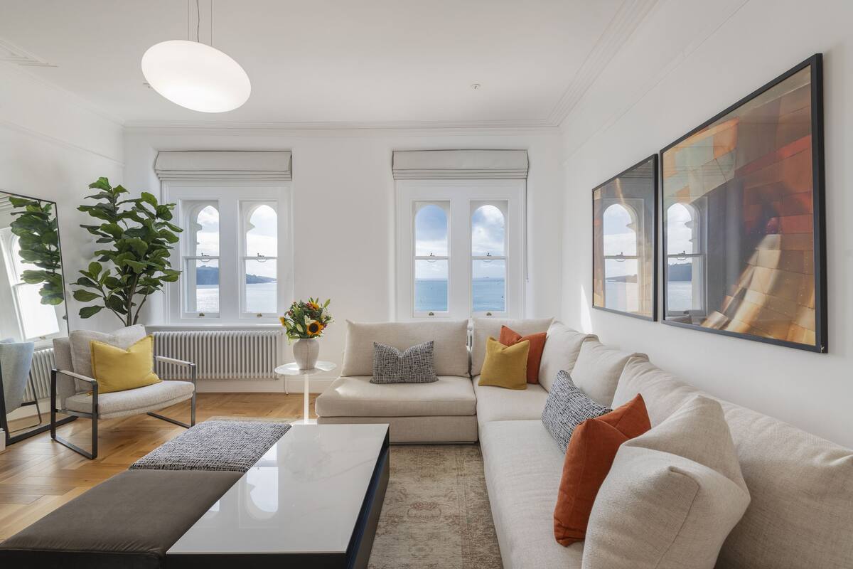 Uma sala de estar moderna com sofá, poltrona, mesa de centro, janelas amplas e vista, e um vaso com planta.