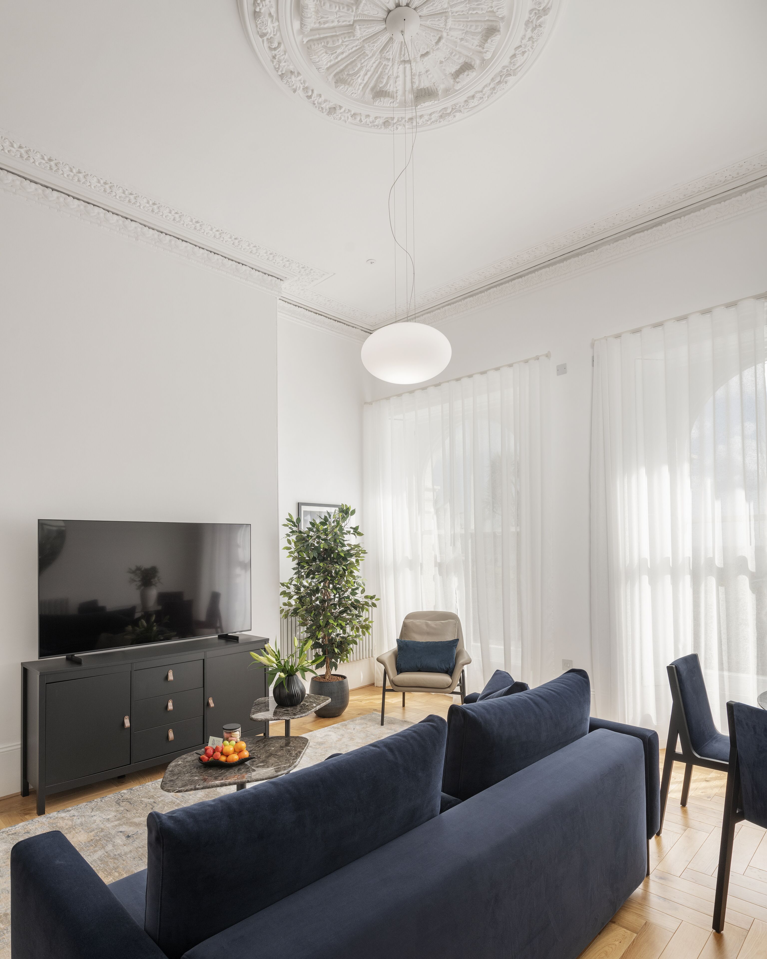 A modern living room with a large flat-screen TV, dark blue sofas, a potted plant, and a large window with sheer curtains.