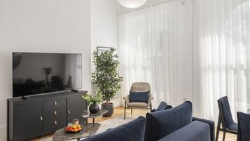 A modern living room with a large flat-screen TV, dark blue sofas, a potted plant, and a large window with sheer curtains.