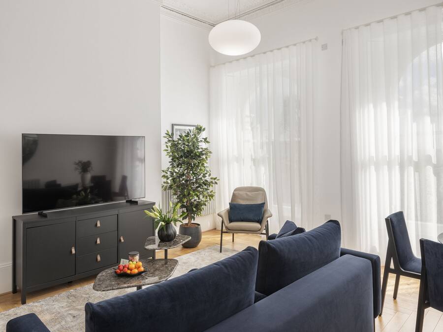 A modern living room with a large flat-screen TV, dark blue sofas, a potted plant, and a large window with sheer curtains.