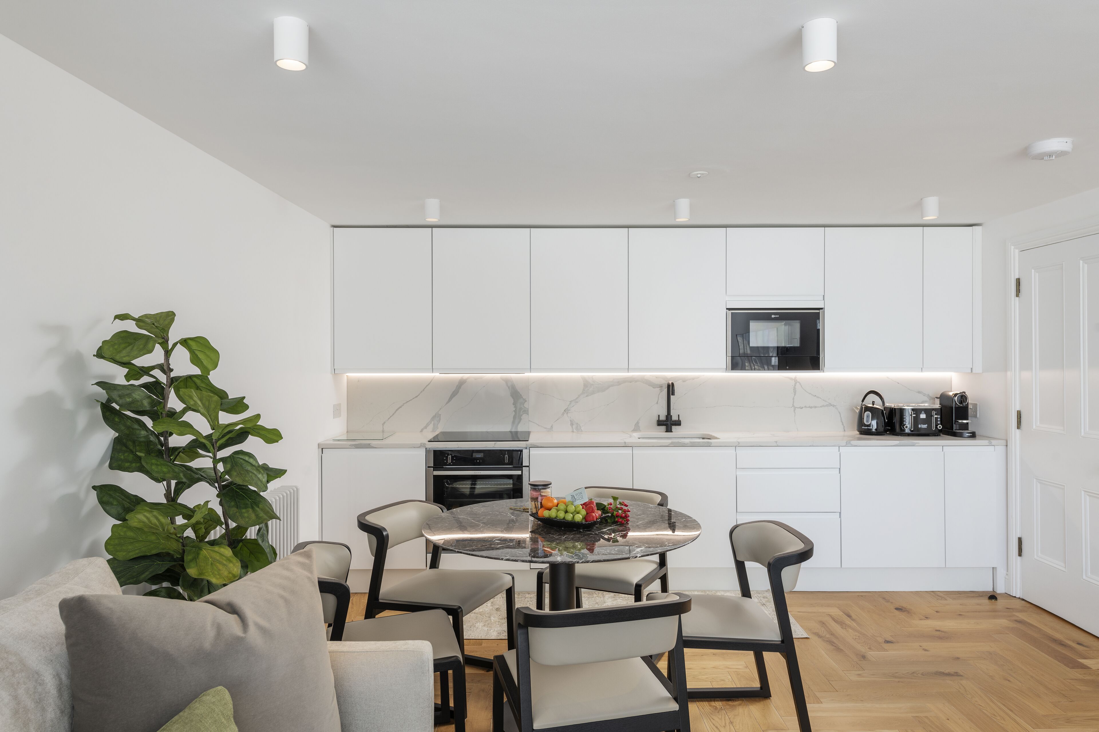 A modern kitchen with white cabinets, a marble countertop, and a dining area with a glass table and chairs.