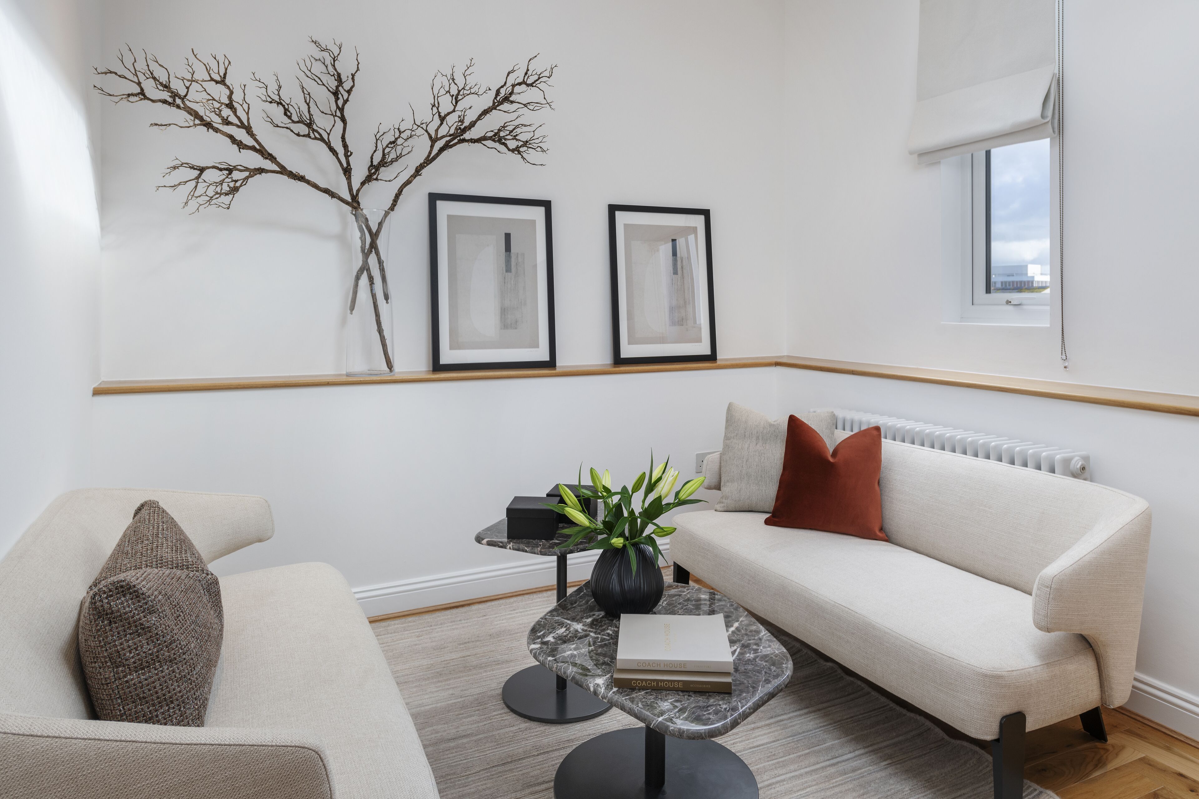 Una sala de estar moderna con un sofá, un sillón y una mesa de centro. Hay dos cuadros en la pared, una planta sobre la mesa de centro y una ventana con vistas.