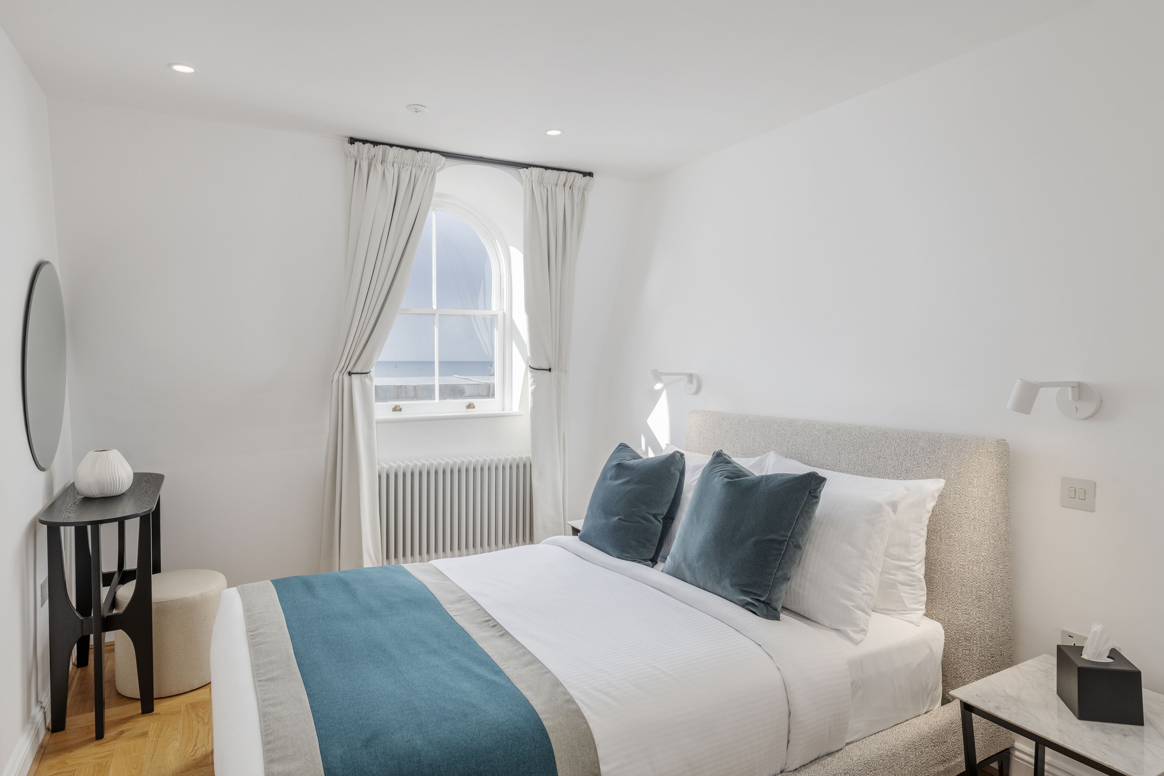 A modern hotel room with a large bed, a bedside table, a small round table, and a window with curtains.