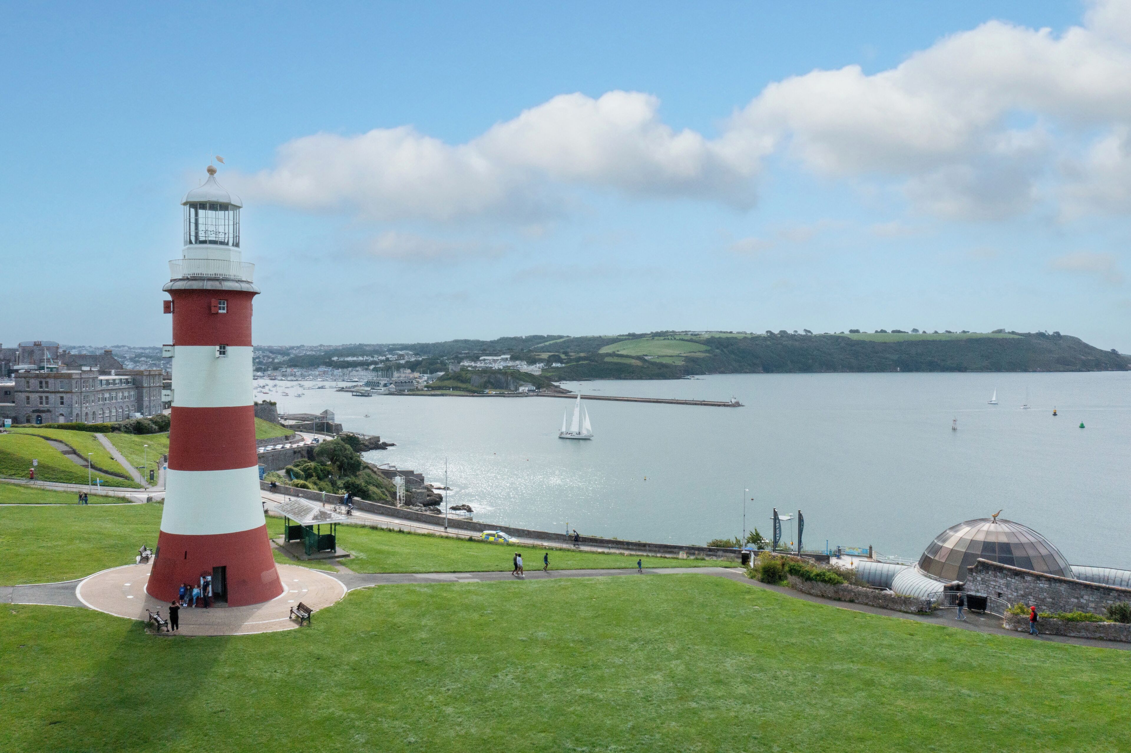 A lighthouse with a red and white striped tower, a grassy area with benches, and a view of the sea with boats.