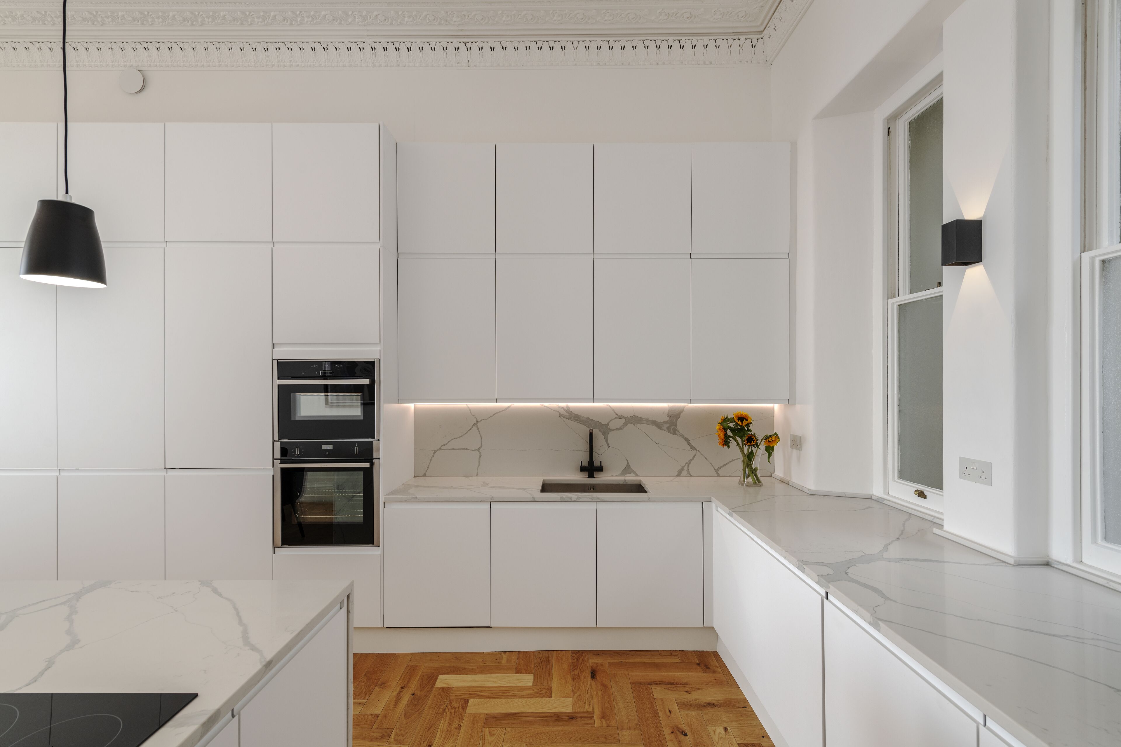 A modern kitchen with white cabinetry, a marble countertop, a built-in oven, and a wooden herringbone floor.