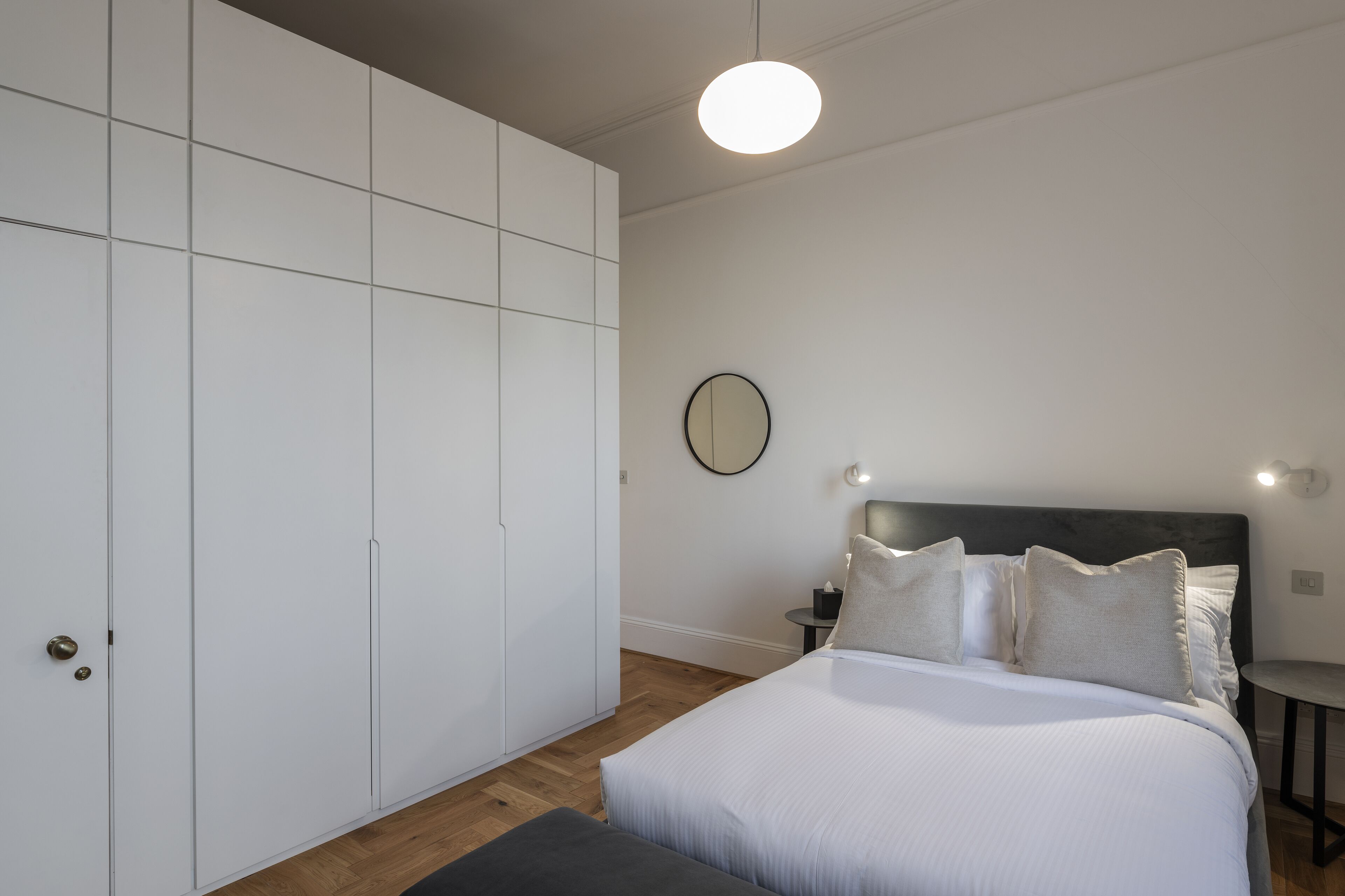 Ein Schlafzimmer mit großem Kleiderschrank, einem Bett mit Kissen, einem runden Spiegel und einem Nachttisch.