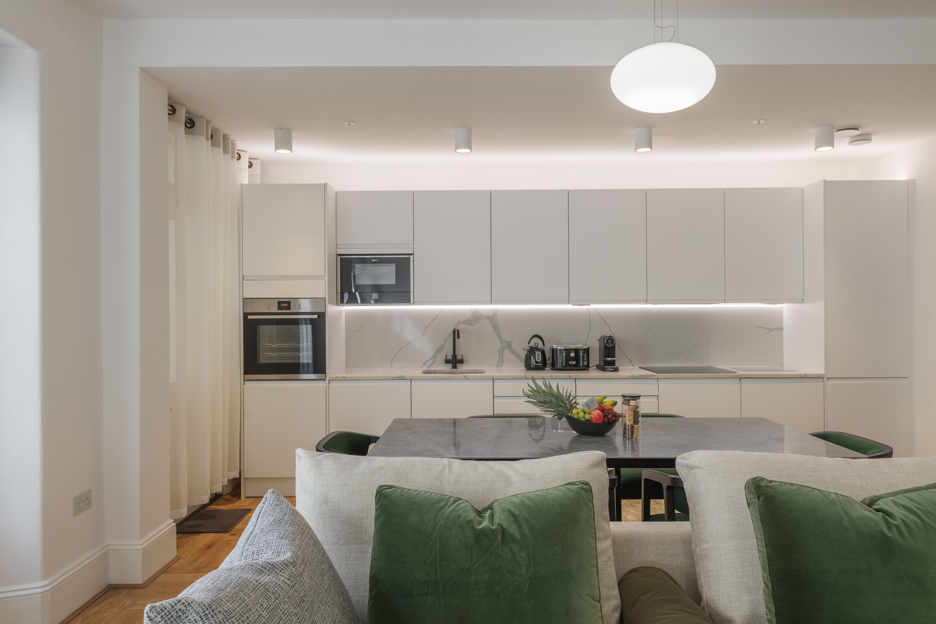 Una cocina moderna con armarios blancos, horno integrado y comedor con mesa y sillas.
