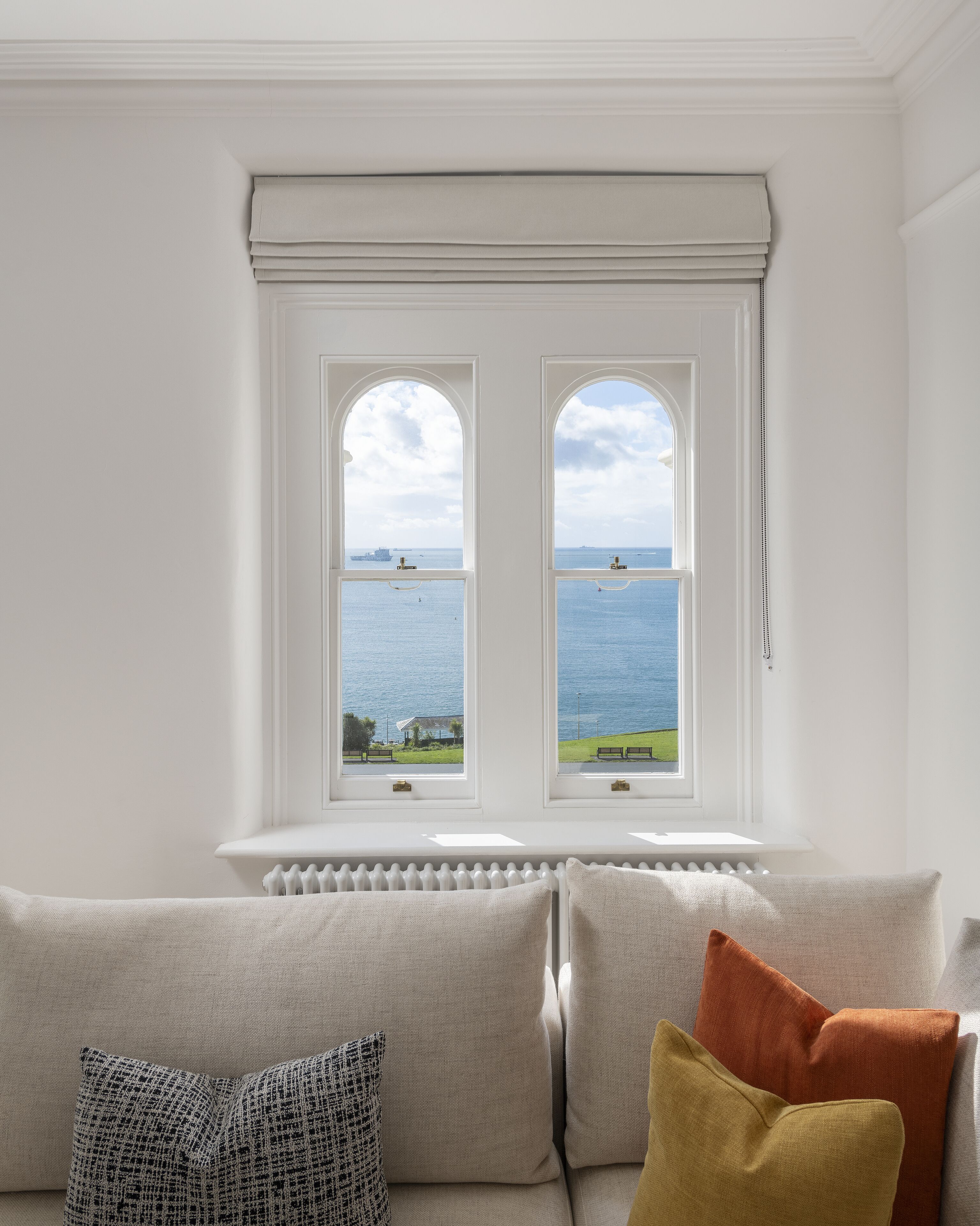 A room with a view of the sea through two windows, a radiator below, and a sofa with cushions.