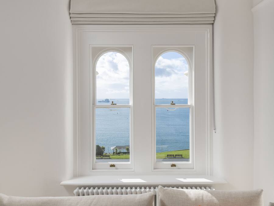 A room with a view of the sea through two windows, a radiator below, and a sofa with cushions.