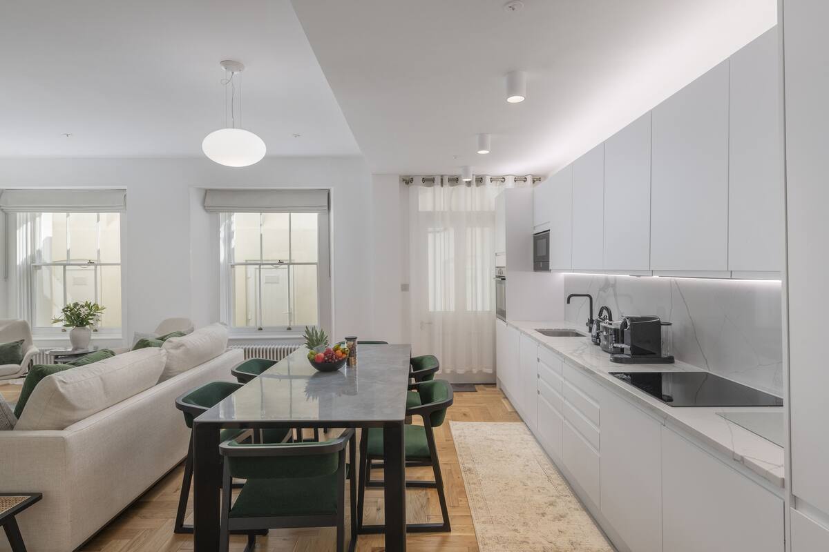 Cozinha moderna com armários brancos, área de jantar com mesa de vidro e sala de estar com sofá e poltronas.