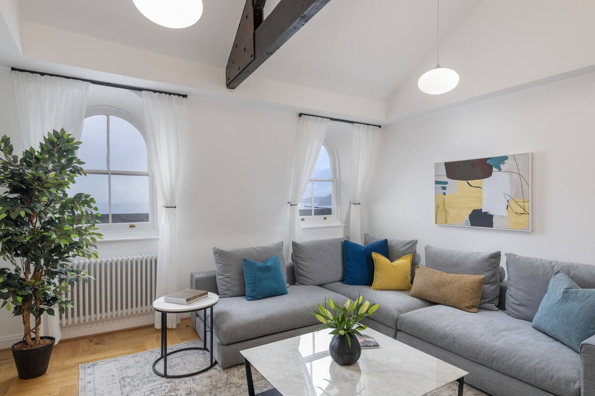Uma sala de estar moderna com um sofá cinza, uma mesinha, uma planta em vaso e uma janela com cortinas.