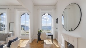 Una habitación amplia con suelo de madera, chimenea, escritorio con silla y grandes ventanales de arco que ofrecen vista al mar.