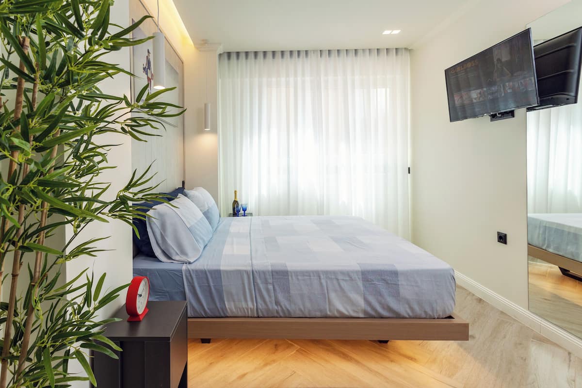 A modern bedroom with a bed, a bedside table with a clock, a TV mounted on the wall, and a large window with sheer curtains.