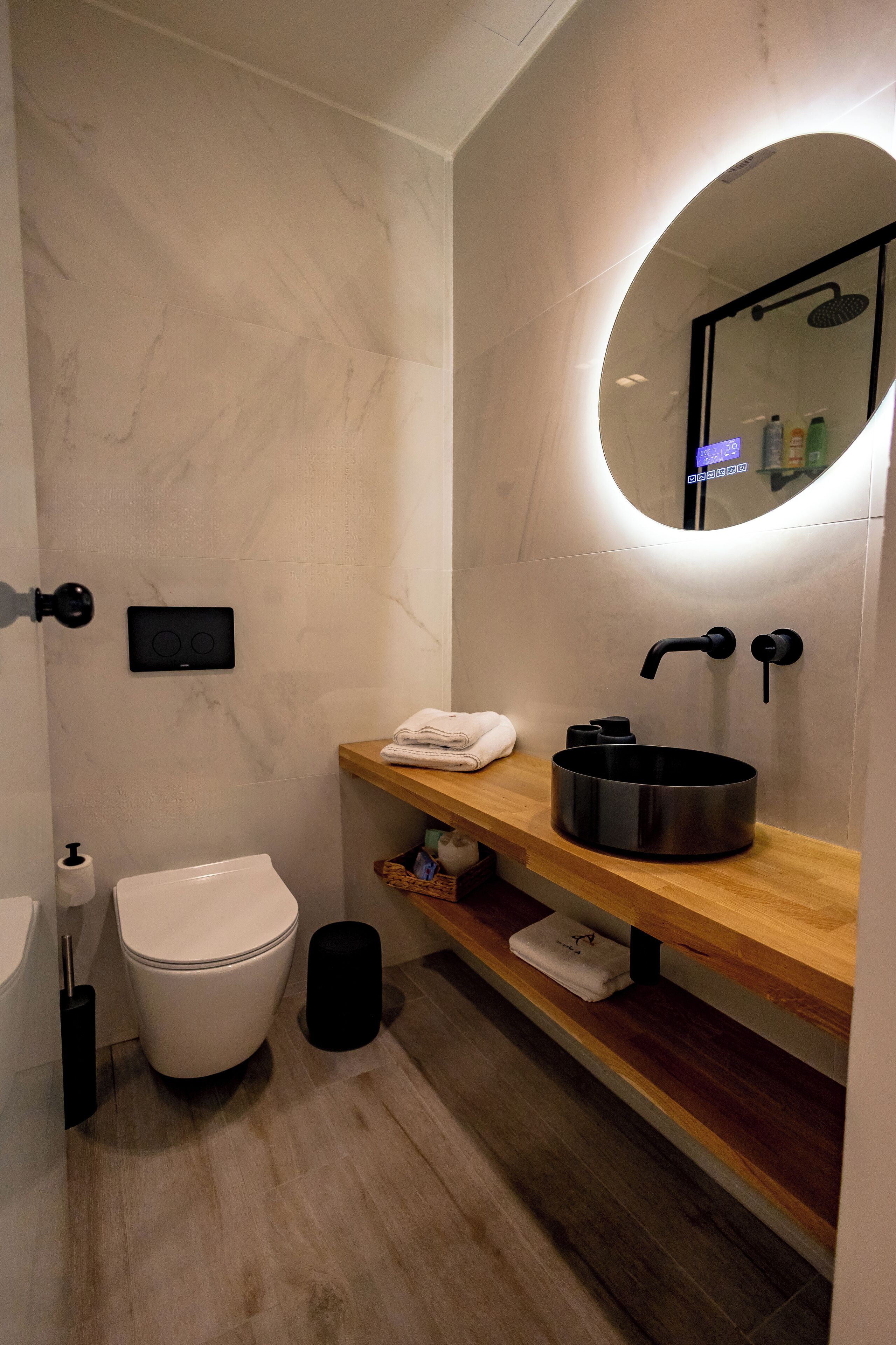 A modern bathroom with a marble wall, a wooden vanity, a black sink, a toilet, and a mirror.