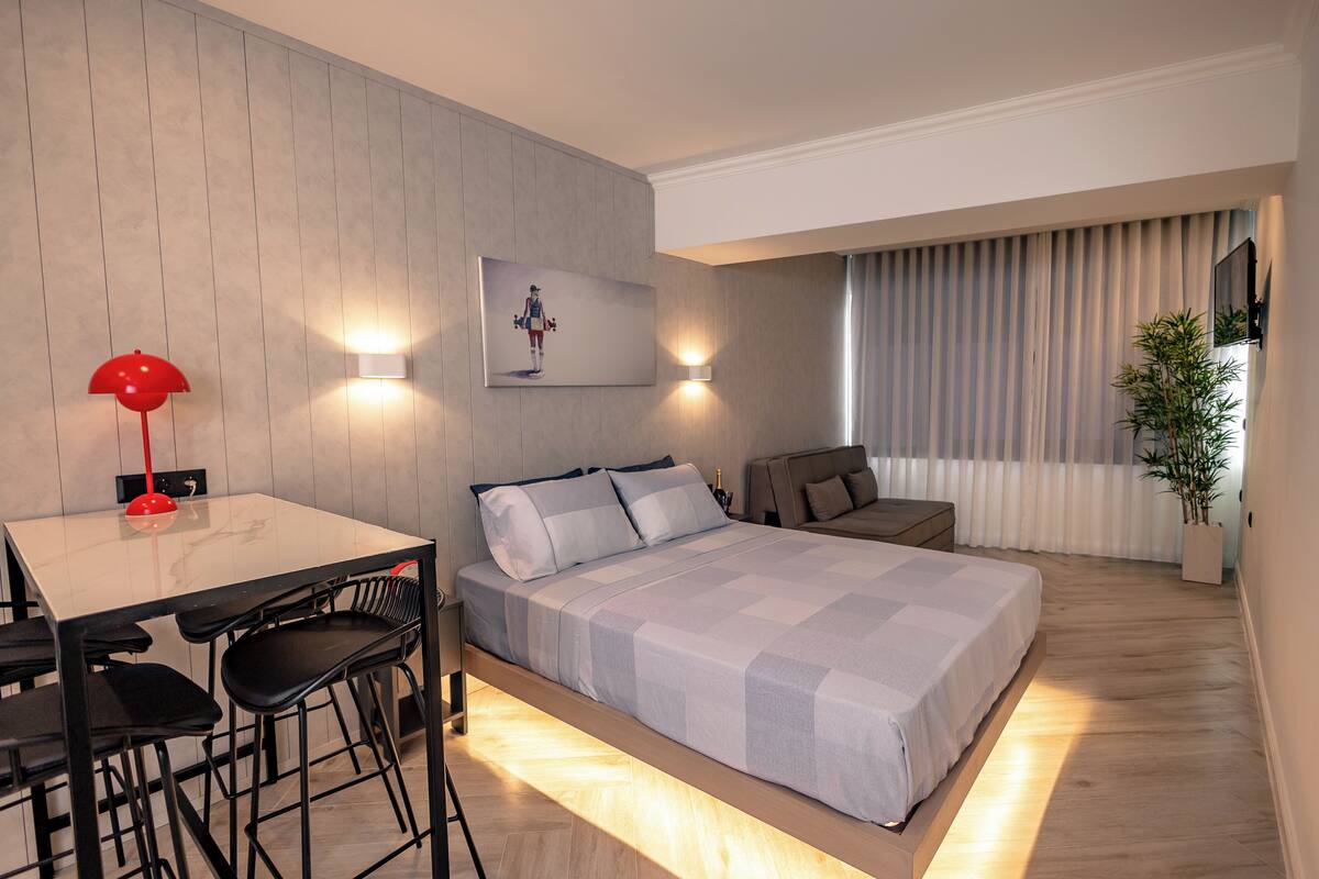 A modern hotel room with a bed, a desk with a chair, a red lamp, and a wall-mounted light.