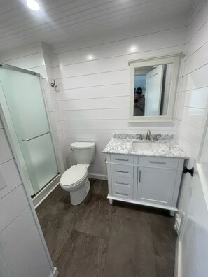A bathroom with a glass shower enclosure, a toilet, a sink with a marble countertop, and a mirror above the sink.
