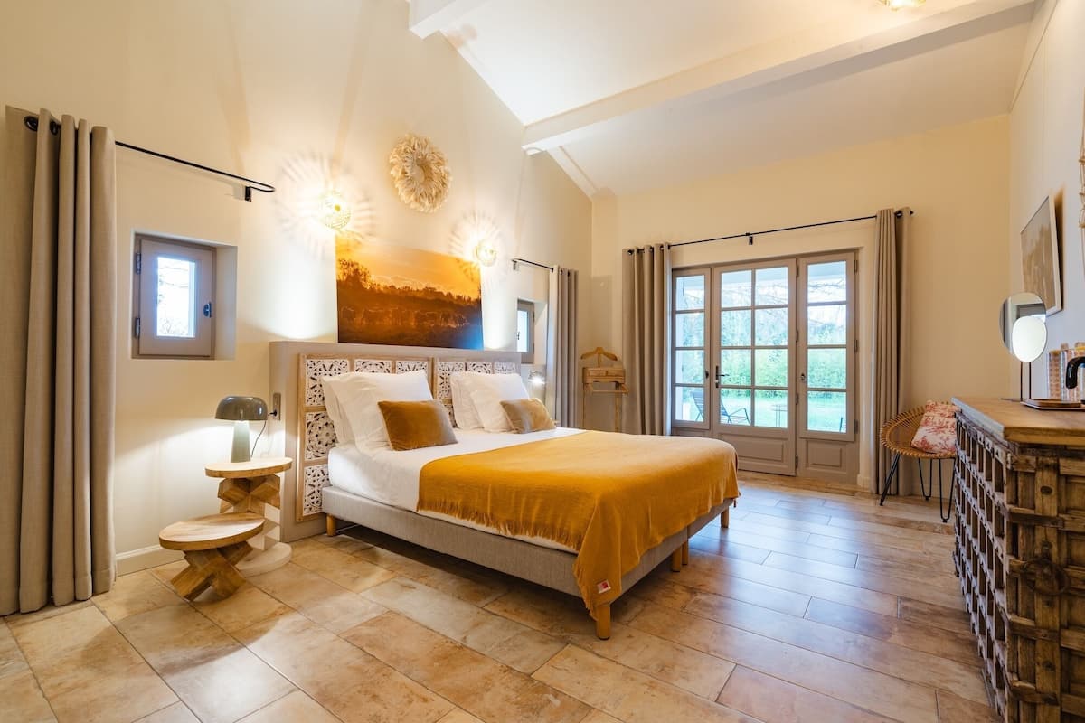 Une chambre spacieuse avec un grand lit, une petite table de chevet, un miroir et une vue sur l’extérieur grâce à une grande fenêtre.