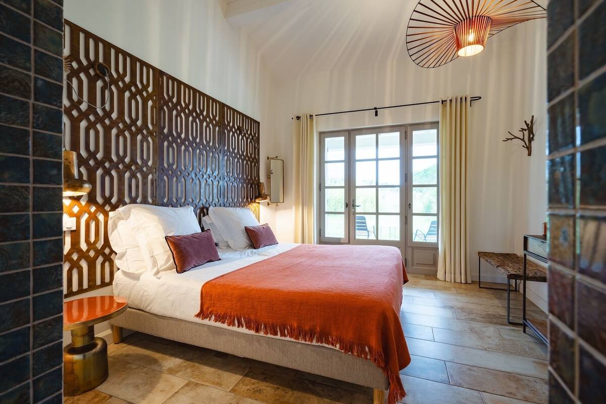 Une chambre à coucher avec un grand lit, un panneau décoratif au mur, une table de chevet, un luminaire suspendu et une fenêtre avec des rideaux.