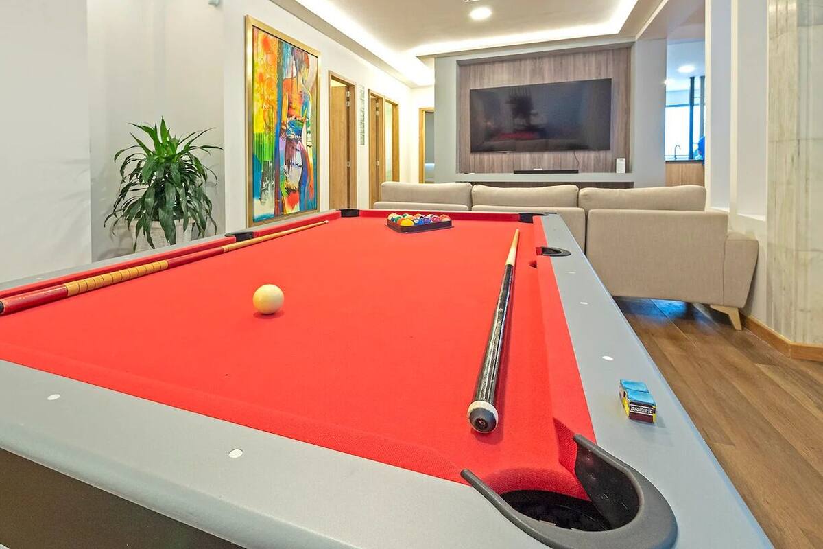 Ein Billardtisch mit roter Tischdecke, eine weiße Billardkugel und ein Billardqueue. Links befindet sich ein Blumentopf und im Hintergrund ein Fernseher.