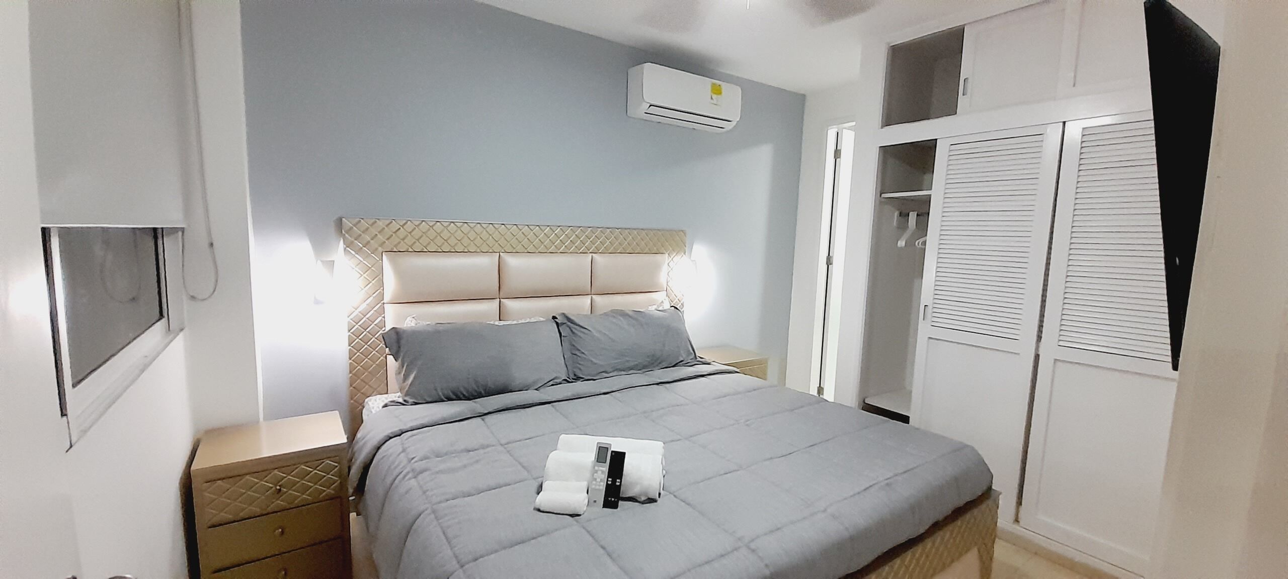 Ein ordentlich bezogenes Bett mit einer grauen Decke, ein Nachttisch mit Lampe und ein Kleiderschrank mit Schiebetüren.
