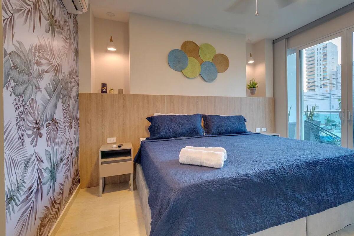 Ein modernes Schlafzimmer mit einem großen Bett, einem Nachttisch, einer Wandleuchte und Blick auf die Stadt durch große Fenster.