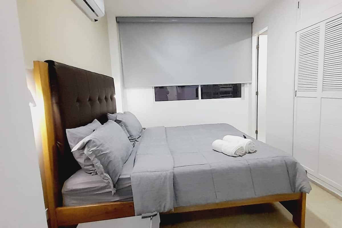 Ein kleines, ordentlich bezogenes Bett mit einer grauen Bettdecke und Kissen, ein hölzernes Kopfstück und ein Fenster mit Jalousien.