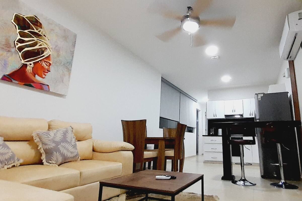 Ein Wohnzimmer mit einer Couch, einem Couchtisch und einem Bild an der Wand. Ein Fernseher ist an der Wand montiert und es gibt einen Deckenventilator.