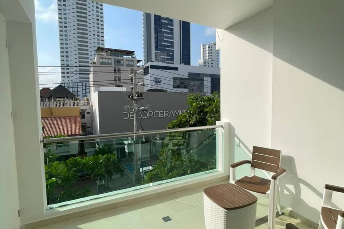 Ein Balkon mit Glasgeländer, weißen Korbstühlen und Blick auf Stadtgebäude.