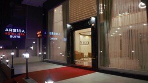 L’extérieur de l’hôtel ARSISA HOTEL-SPA SUITE se distingue par son entrée surmontée d’un tapis rouge, ses grandes fenêtres et son intérieur visible, où l’on aperçoit des espaces aménagés avec des sièges.
