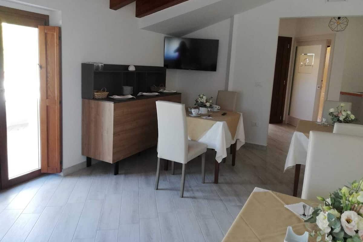 A dining area with a wooden table and chairs, a kitchen counter with a TV, and a door leading to another room.