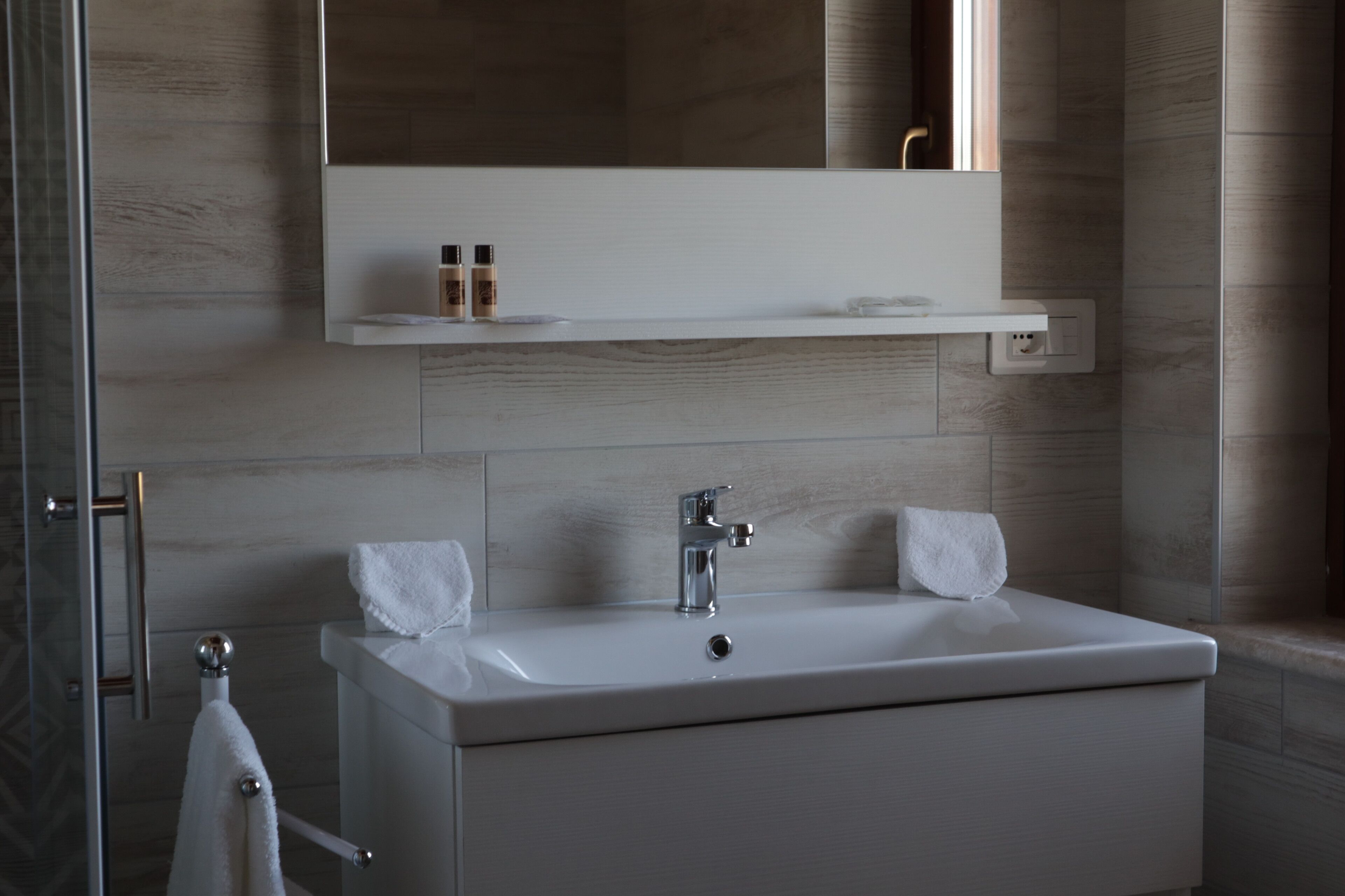 A modern bathroom with a white sink, two towels, a mirror above the sink, and a shelf with toiletries.