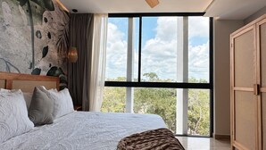 A bedroom with a large bed, a ceiling fan, a view of trees through a window, and a wooden wardrobe. - Alterhome Amour 307 (Tulum)