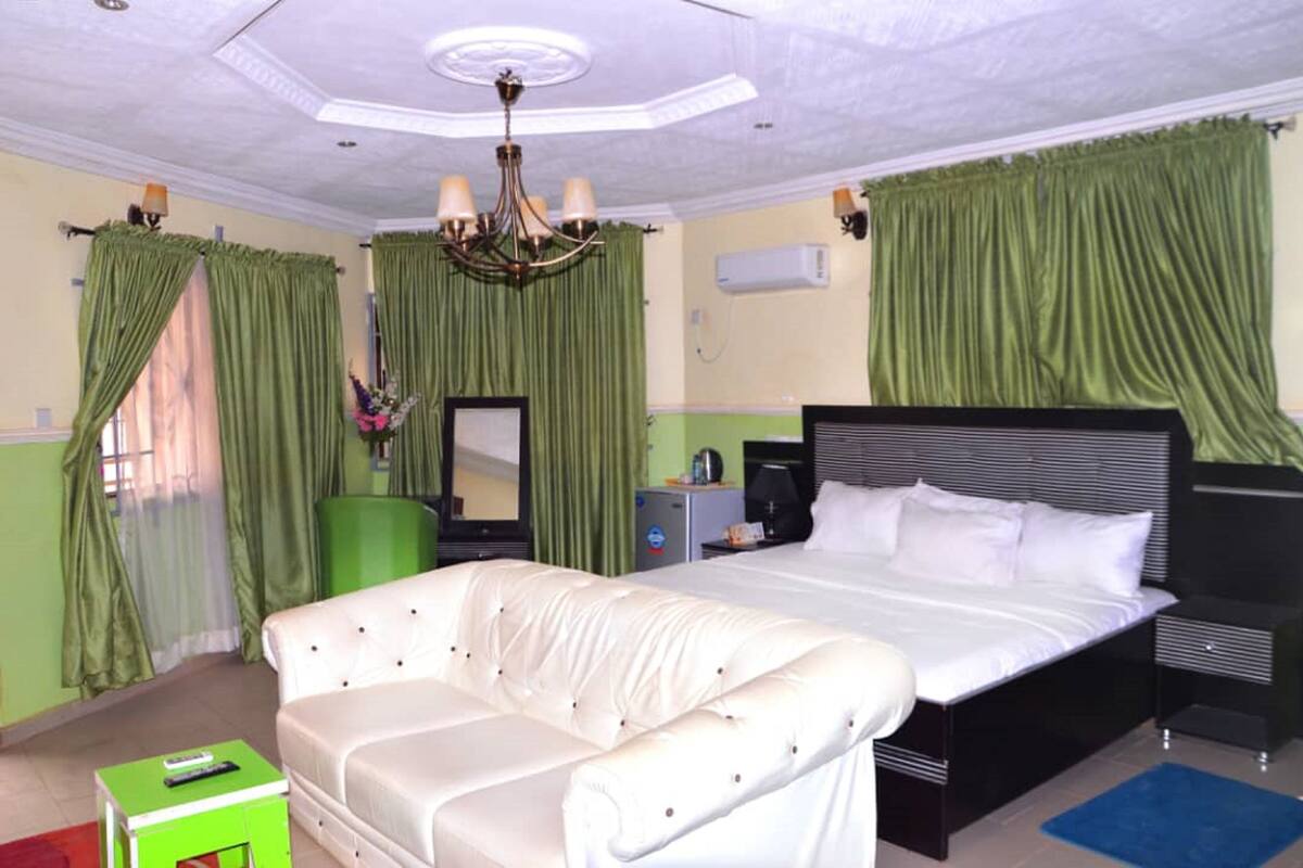 A hotel room with a large bed, a white sofa, a green armchair, a desk with a lamp, and a ceiling fan.