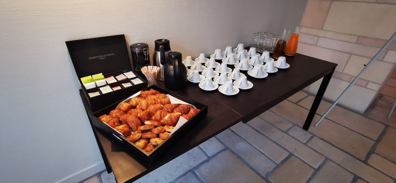 A table with a box of chocolates, a tray of croissants, and several coffee cups and glasses.