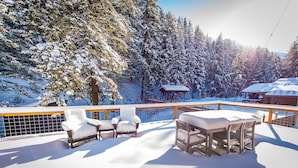 A snow-covered outdoor seating area with a view of a forest and a wooden structure in the background.
