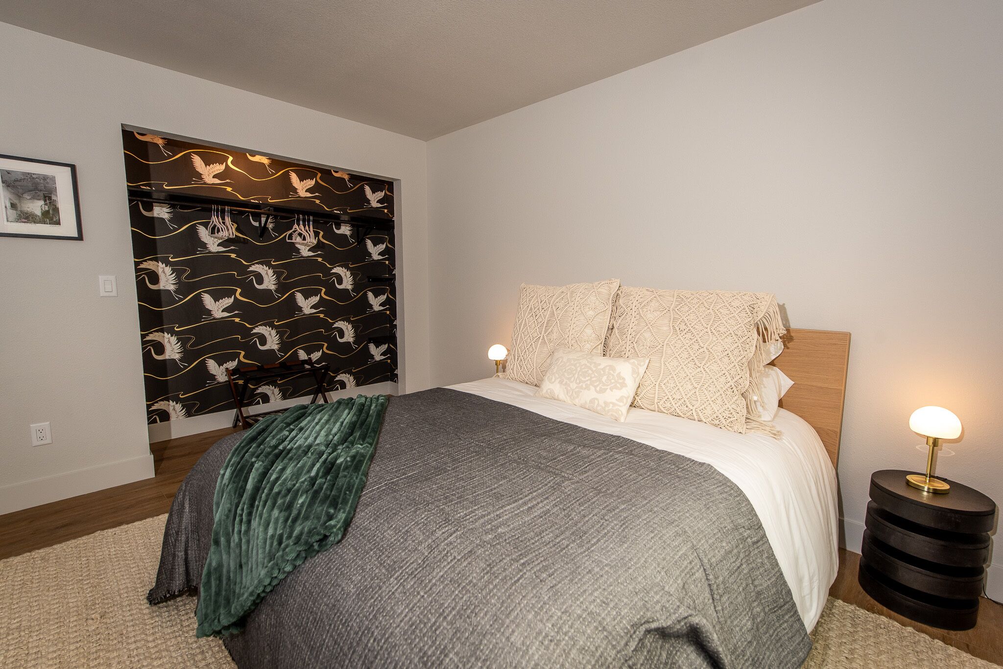 A bedroom with a bed, a nightstand, a wall-mounted shelf with decorative items, and a framed picture on the wall.