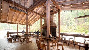 A rustic wooden interior with a stone fireplace, wooden tables and chairs, and large windows overlooking a forested area.