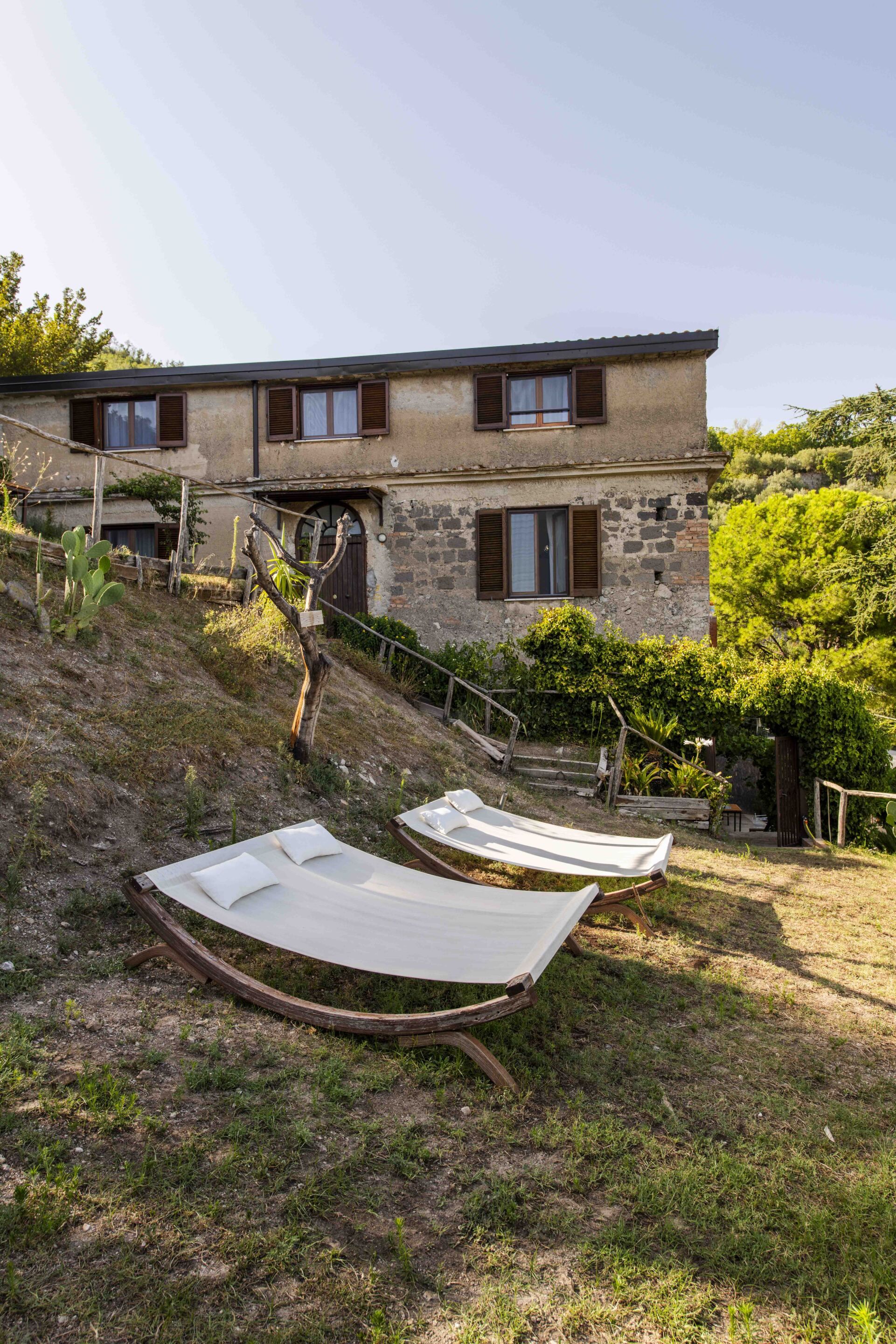 A rustic building with a sloped roof, wooden shutters, and a stone wall. There are two hammocks on the ground in front of the building.