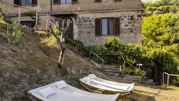 A rustic building with a sloped roof, wooden shutters, and a stone wall. There are two hammocks on the ground in front of the building.