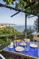 An outdoor dining table with a view of the sea and hills, set with plates, glasses, and a basket of fruit.