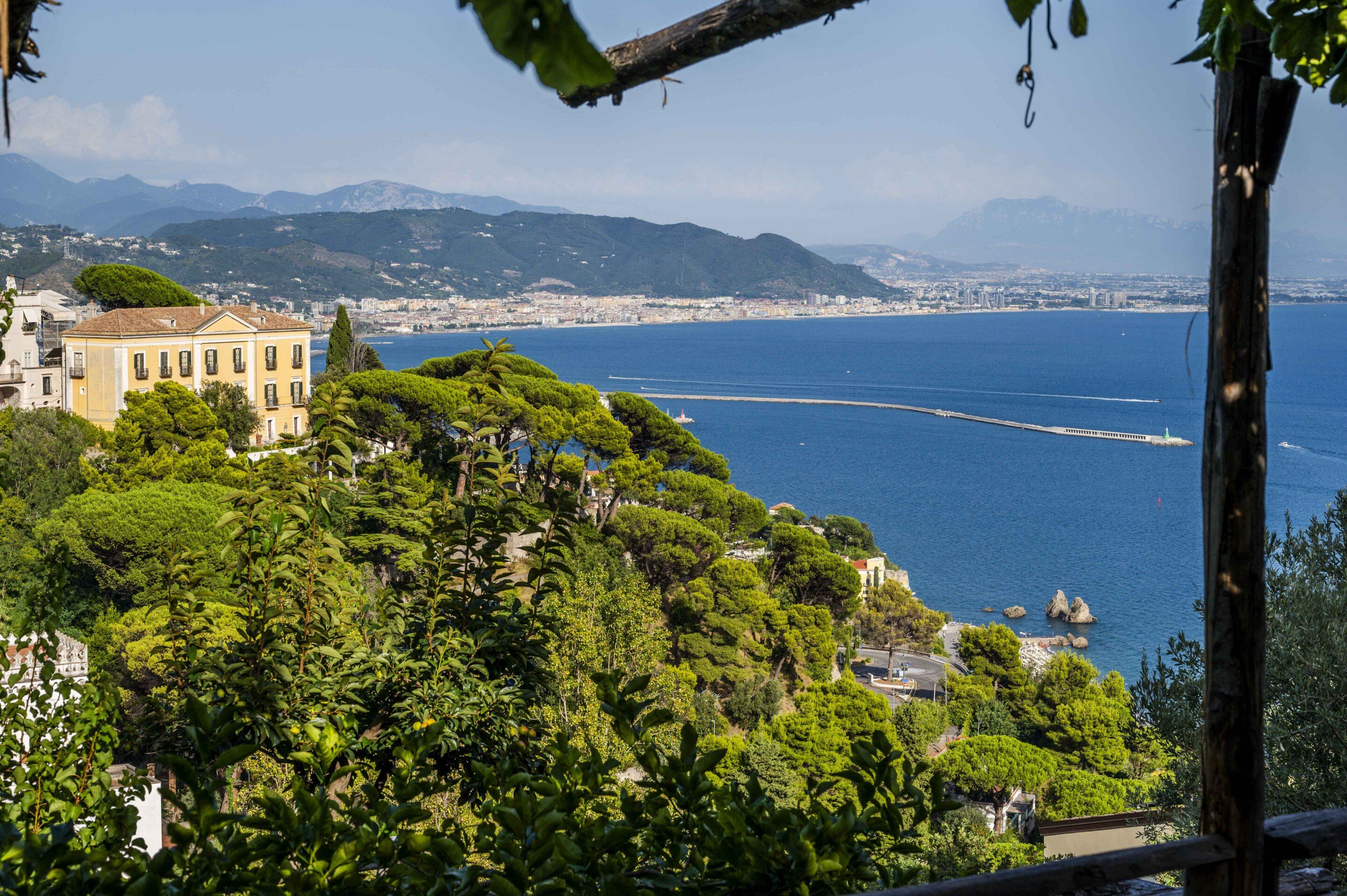 A coastal cityscape with a prominent building overlooking the sea, lush greenery, and distant mountains.