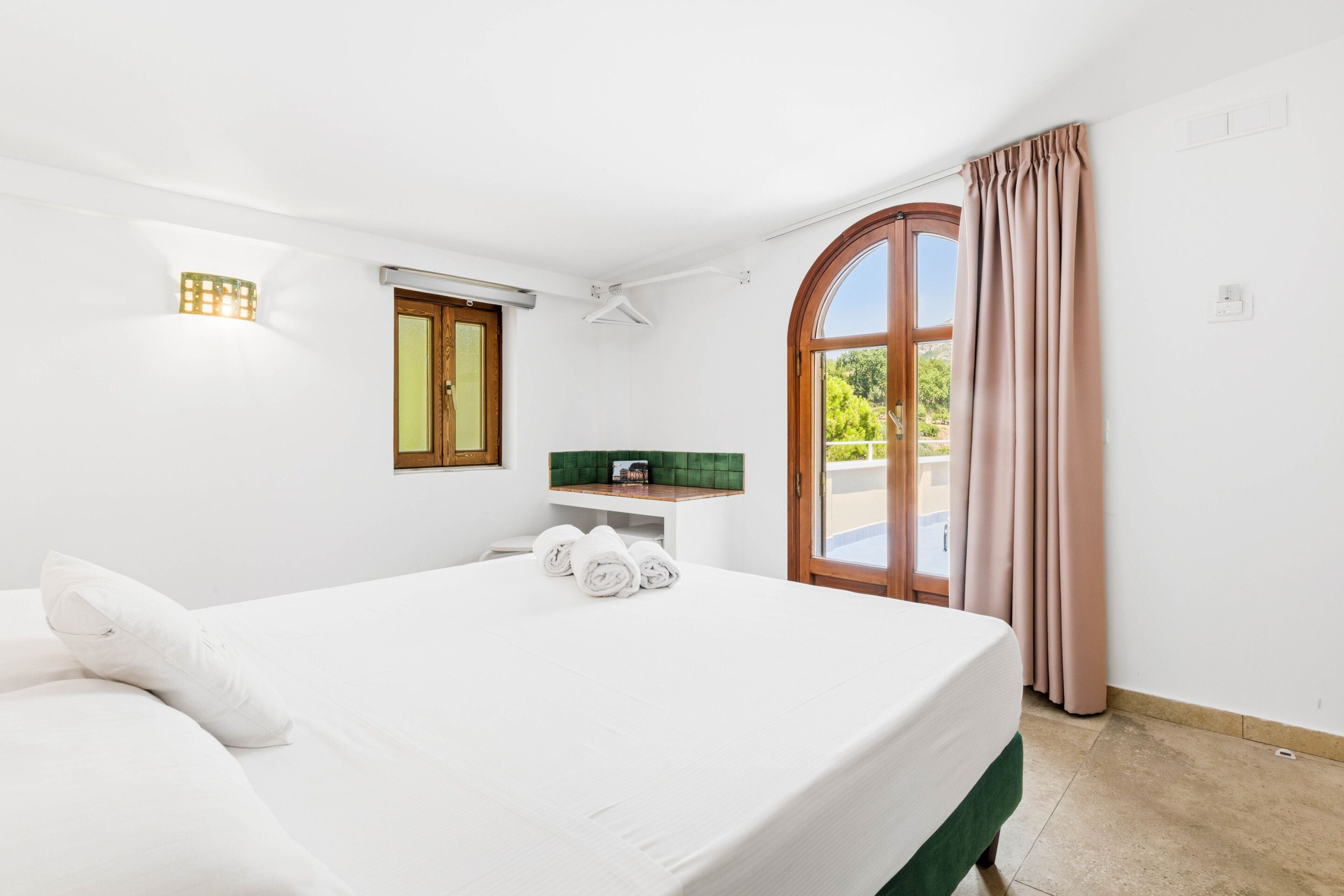 A hotel room with a bed, a small table, a green cushioned bench, a window with curtains, and a view of greenery outside.